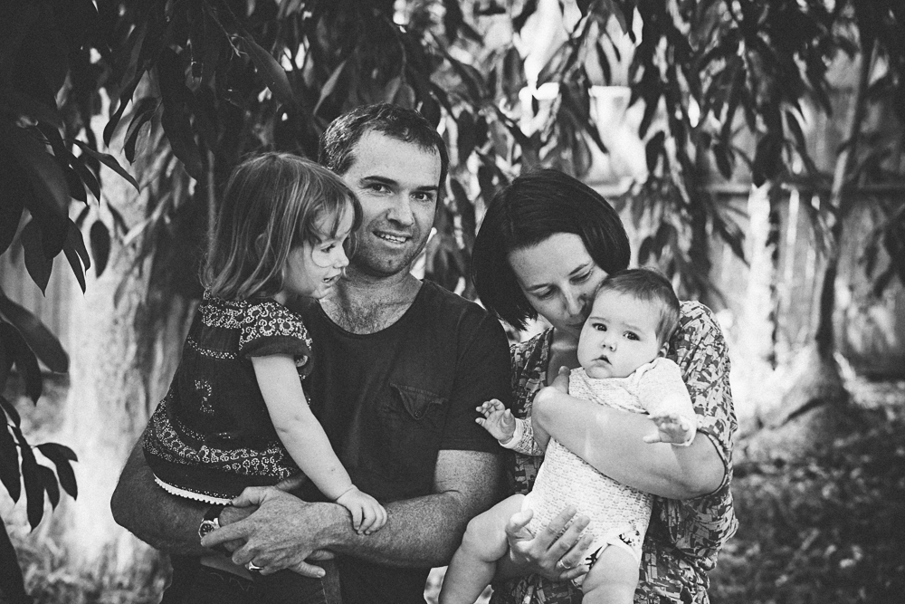 Black and white family portrait, of a white dad holding toddler, mum holding baby, standing under a willow tree. Only the dad is looking to camera