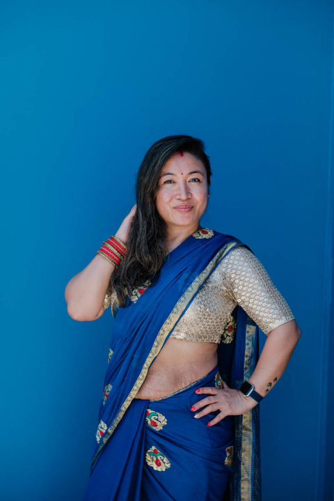 Image of a woman standing against a blue wall with one hand on her hip the other in her hair wearing a blue Nepalese sari. Her beautiful stretchmarks visible on her stomach
