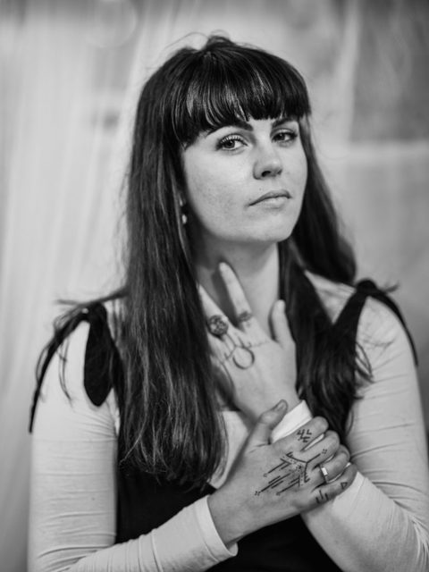 Black and white portrait of a woman with long dark hair, full fringe. She is holding her tattooed hands to her heart and throat, looking into the camera, unsmiling