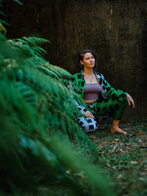 Colour portrait of a woman on her haunches, partially obscured by green fern fronds. Her expression is strong and peaceful. Unsmiling, looking to camera