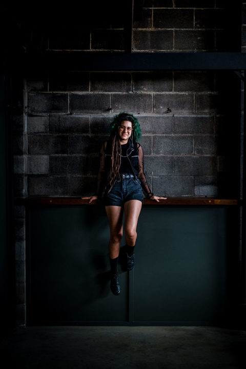 Coloured portrait of a young woman perched on a shelf, against a block wall. She has green hair, dreadlocks, tattoos and is wearing all black